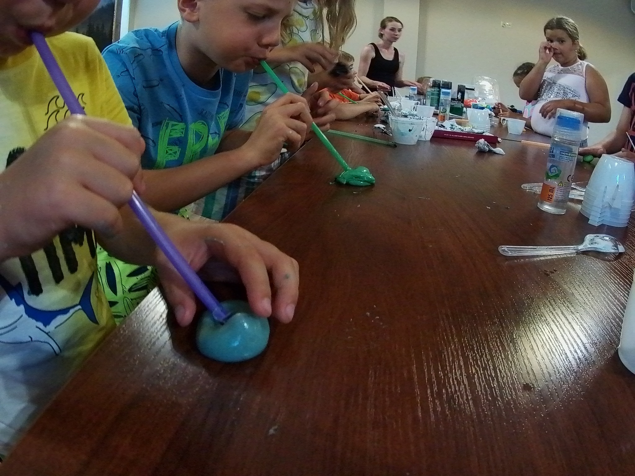Dzieci w Odrowążu podczas warsztatów tworzenia slime, dmuchają przez słomki w kolorowe masy na drewnianym stole, widoczne kubeczki, kleje i inne akcesoria.