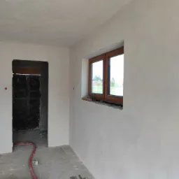 Wnętrze w stanie deweloperskim z oknem i surowymi ścianami po nałożeniu warstwy tynku, widoczne elementy instalacji grzewczej podłogowej i materiały budowlane.