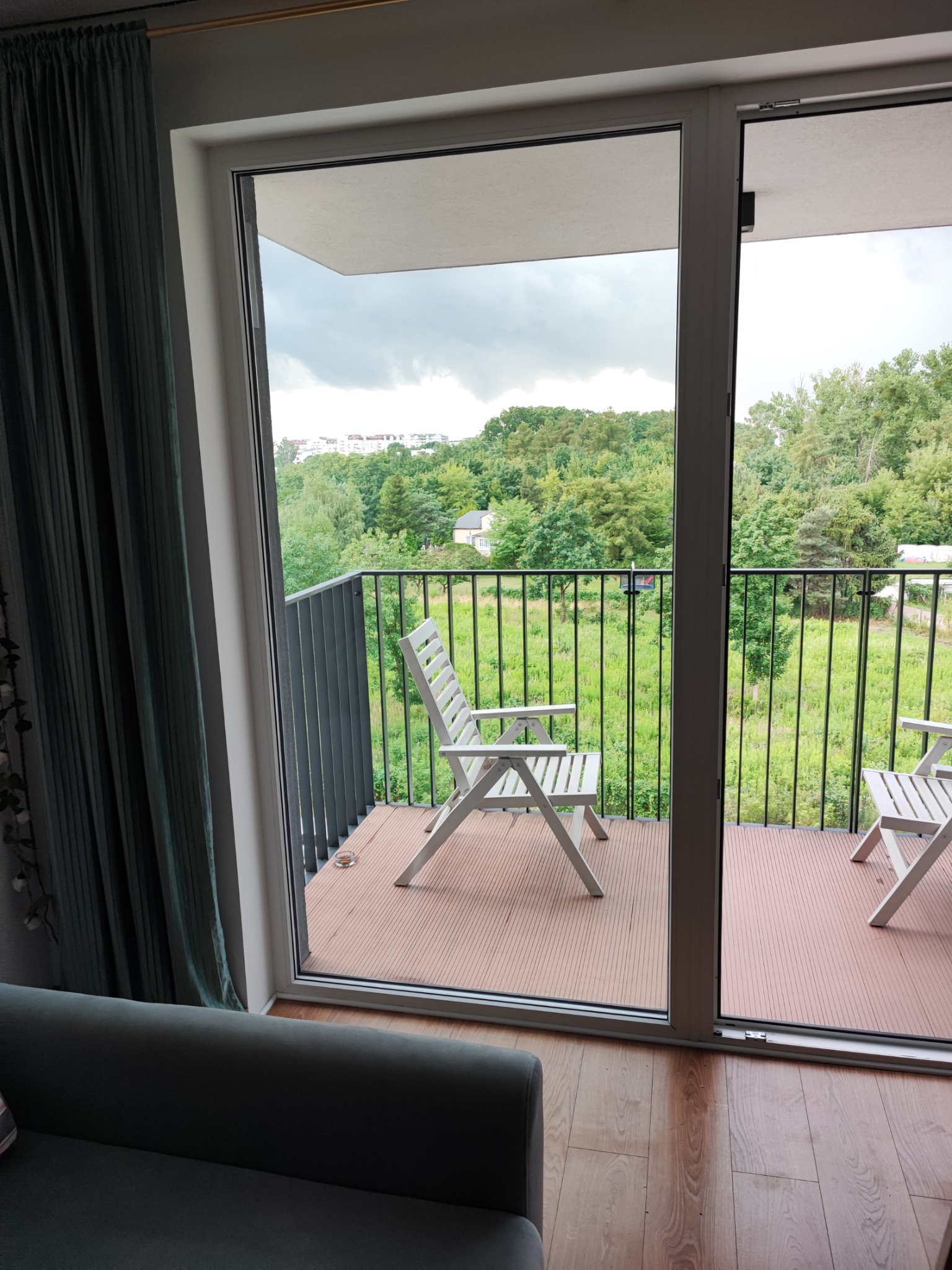 Balkon z czarną metalową balustradą, dwoma białymi krzesłami i brązową podłogą z widokiem na zieleń i budynki w tle, widziany z wnętrza mieszkania przez otwarte drzwi balkonowe.