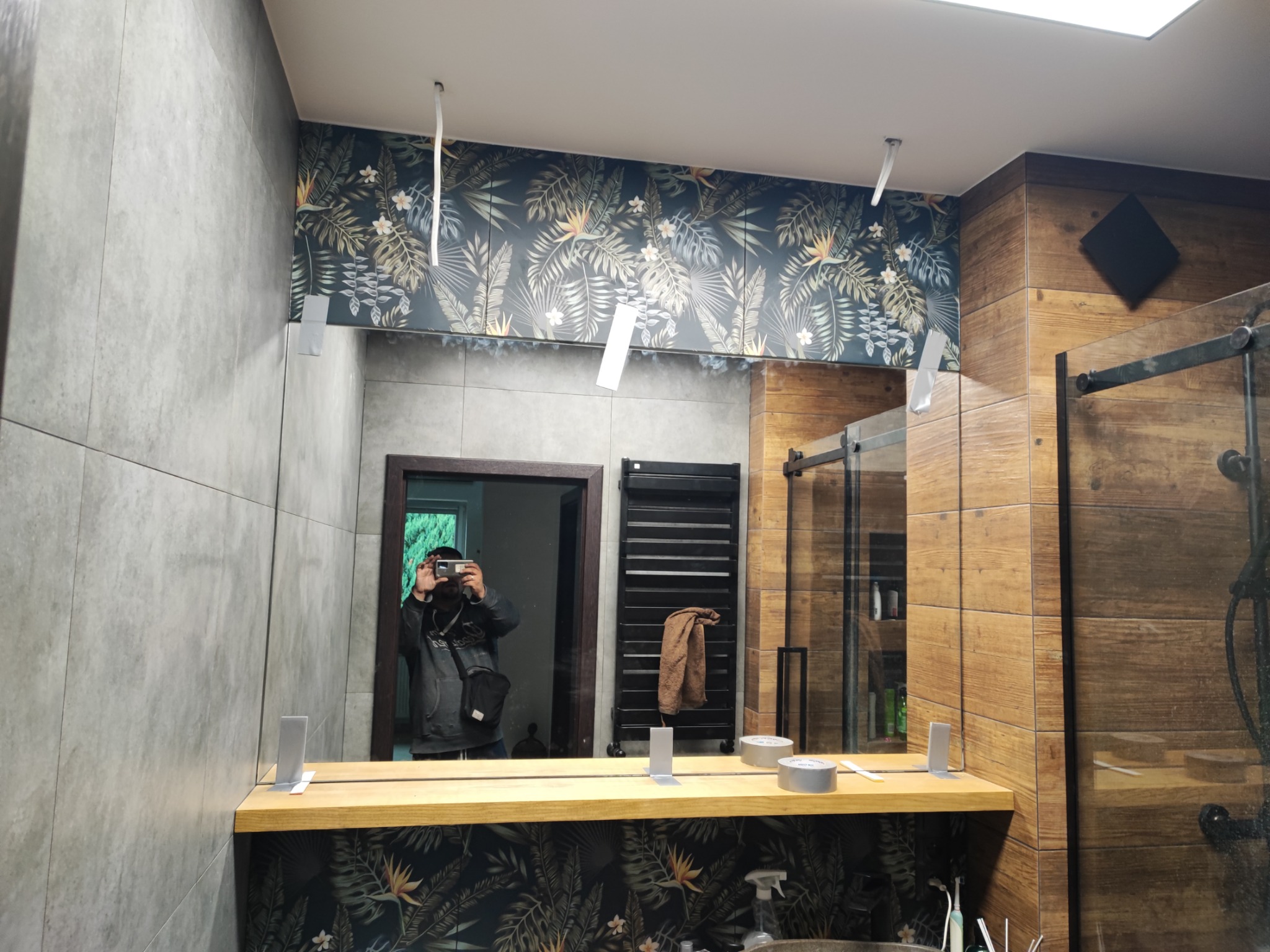 Nowoczesna łazienka z drewnianymi i szarymi płytkami, ozdobiona tapetą w tropikalne liście; widoczne lustro z drewnianym blatem i osoba robiąca zdjęcie.