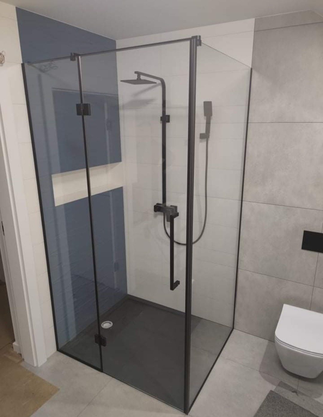 Nowoczesna kabina prysznicowa z czarnymi profilami i deszczownicą, szary brodzik, widoczna toaleta w tle.