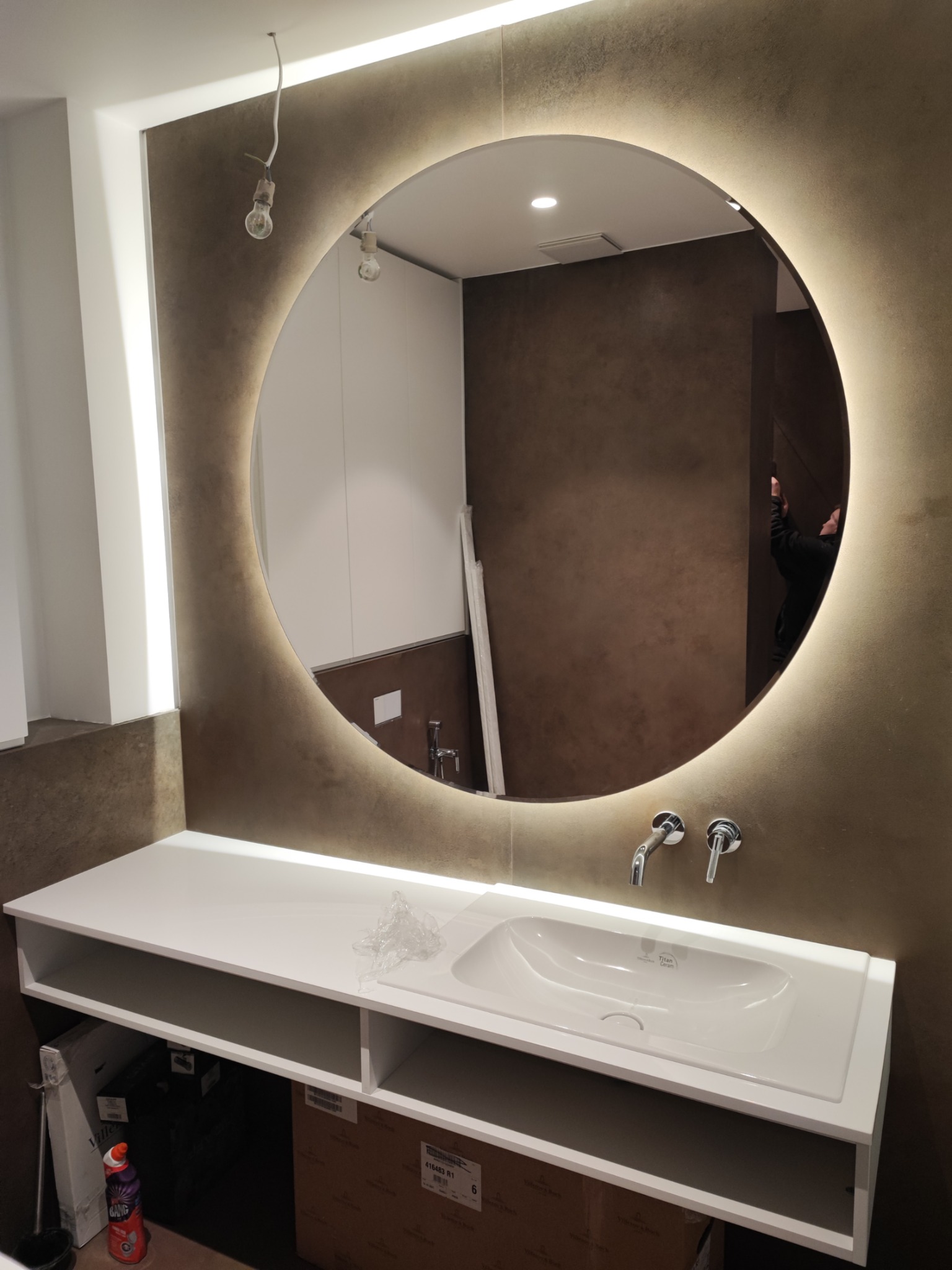 Okrągłe lustro z podświetleniem LED nad białą umywalką zamontowaną na szafce, widoczne niedokończone prace instalacyjne.