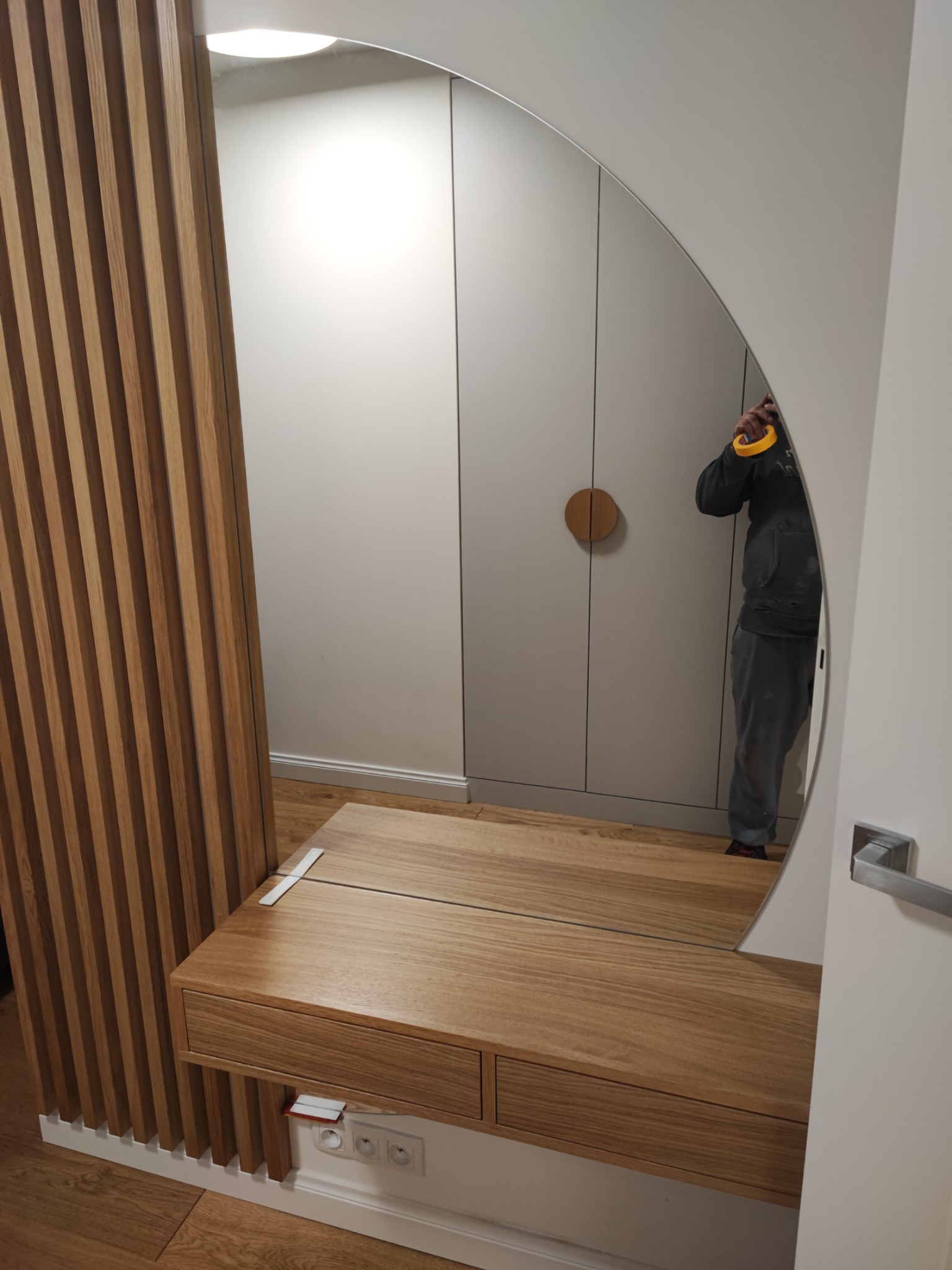 Nowoczesna toaletka z drewnianym blatem i lamelami, z dużym okrągłym lustrem, w którym odbija się osoba z żółtą taśmą malarską.