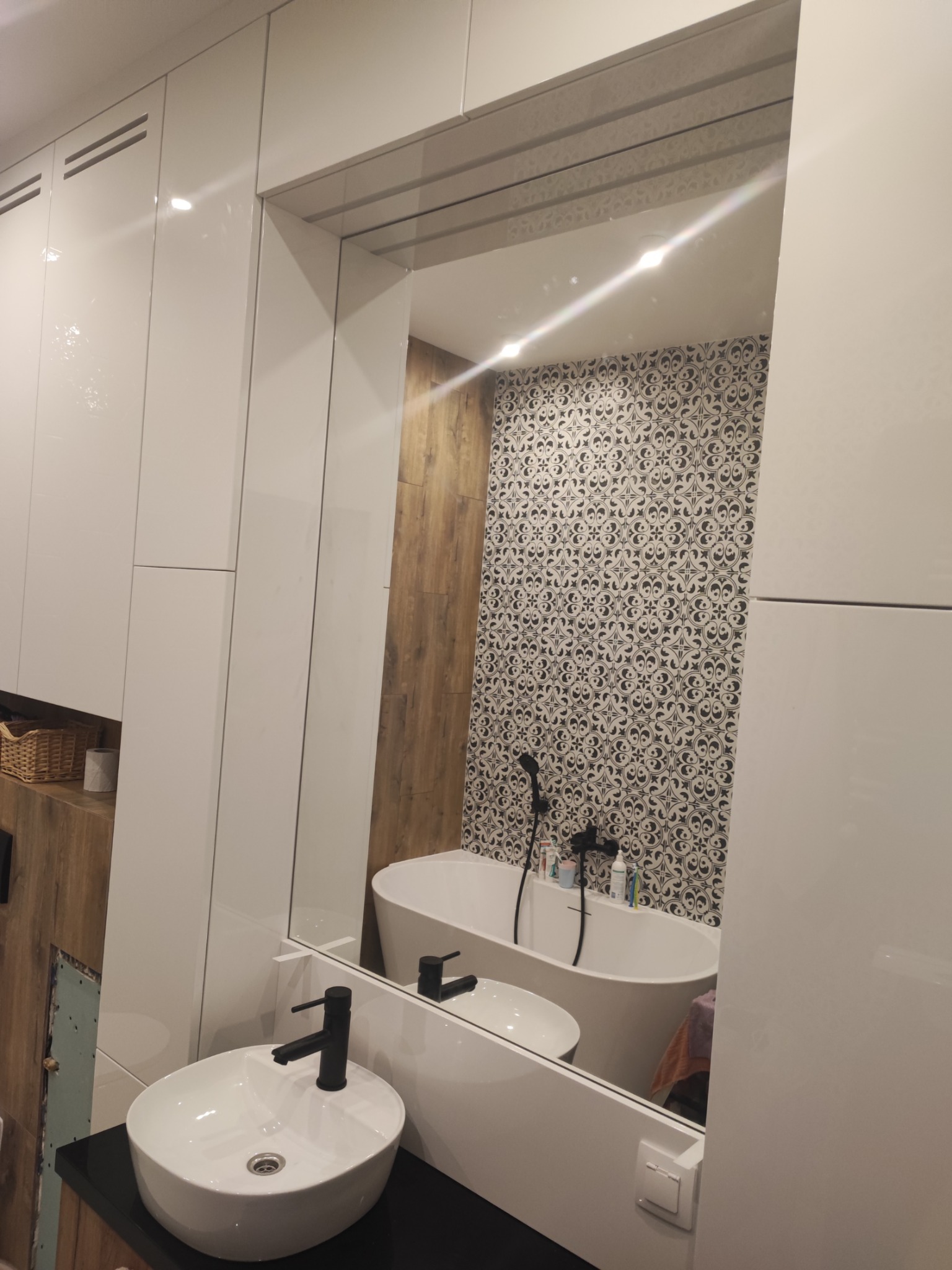 Nowoczesna łazienka z białą armaturą, widoczna w lustrze: owalna umywalka z czarną baterią i wolnostojąca wanna na tle wzorzystych płytek.