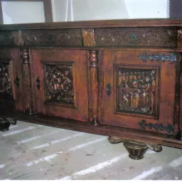 Starodawna, ciemnobrązowa komoda z rzeźbionymi scenami figuralnymi na drzwiach, wsparta na ozdobnych nóżkach w kształcie miseczek, ustawiona na podłodze z widocznymi śladami malowania.