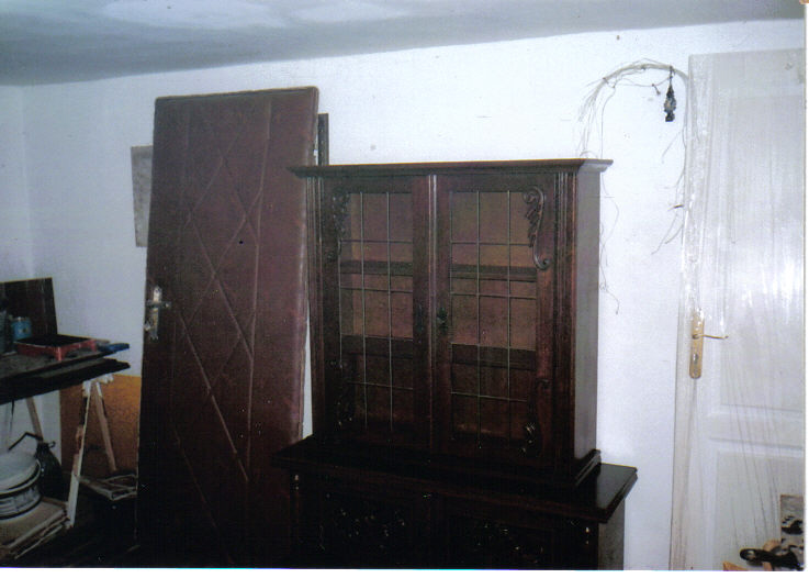 Niedokończony remont pokoju: ciemnobrązowa, lakierowana szafka z przeszklonymi drzwiczkami stoi obok brązowych, wzorzystych drzwi opartych o ścianę, na ścianie wisi dekoracja z gałązek