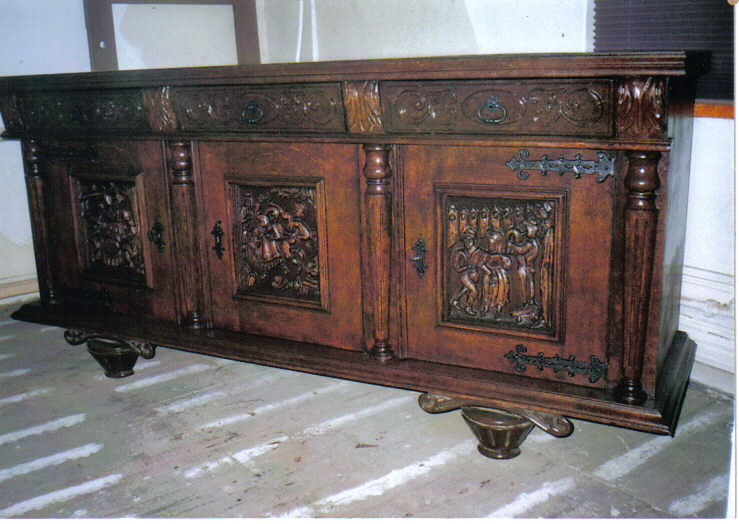 Starodawna, ciemnobrązowa komoda z rzeźbionymi scenami figuralnymi na drzwiach, wsparta na ozdobnych nóżkach w kształcie miseczek, ustawiona na podłodze z widocznymi śladami malowania.