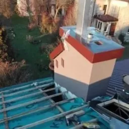 Montaż membrany dachowej z widocznym kominem obłożonym blachą, częściowo pokrytym dachem z czerwonej dachówki, narzędzia dekarskie na membranie, w tle budynki mieszkalne i trawnik.