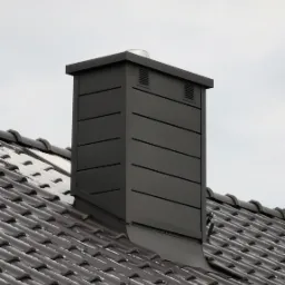 Nowoczesny, szary komin z blachy na dachu pokrytym brązową dachówką, lekko ośnieżonym, na tle pochmurnego nieba.
