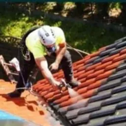 Pracownik w kasku i masce ochronnej, malujący dachówkę na dachu za pomocą pistoletu natryskowego. Część dachu pomalowana na czerwono, pozostała część w kolorze grafitowym. Na drugim planie widoczne...