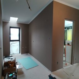 Krystian Wierkin - Pokój w trakcie remontu, ściany pomalowane na brązowo, widoczne narzędzia, płyta gipsowa na podłodze, listwy przypodłogowe i sufitowe zamontowane, instalacja klimatyzacji pod sufitem.