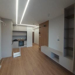 Krystian Wierkin - Nowoczesne wnętrze mieszkania z drewnianą podłogą, oświetleniem LED w suficie i zabudową kuchenną w odcieniach szarości, widoczny fragment kartonu na podłodze.