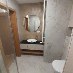 Nowoczesna łazienka z marmurowymi płytkami, okrągłym lustrem, białą umywalką nablatową na czarnym blacie i grzejnikiem drabinkowym.