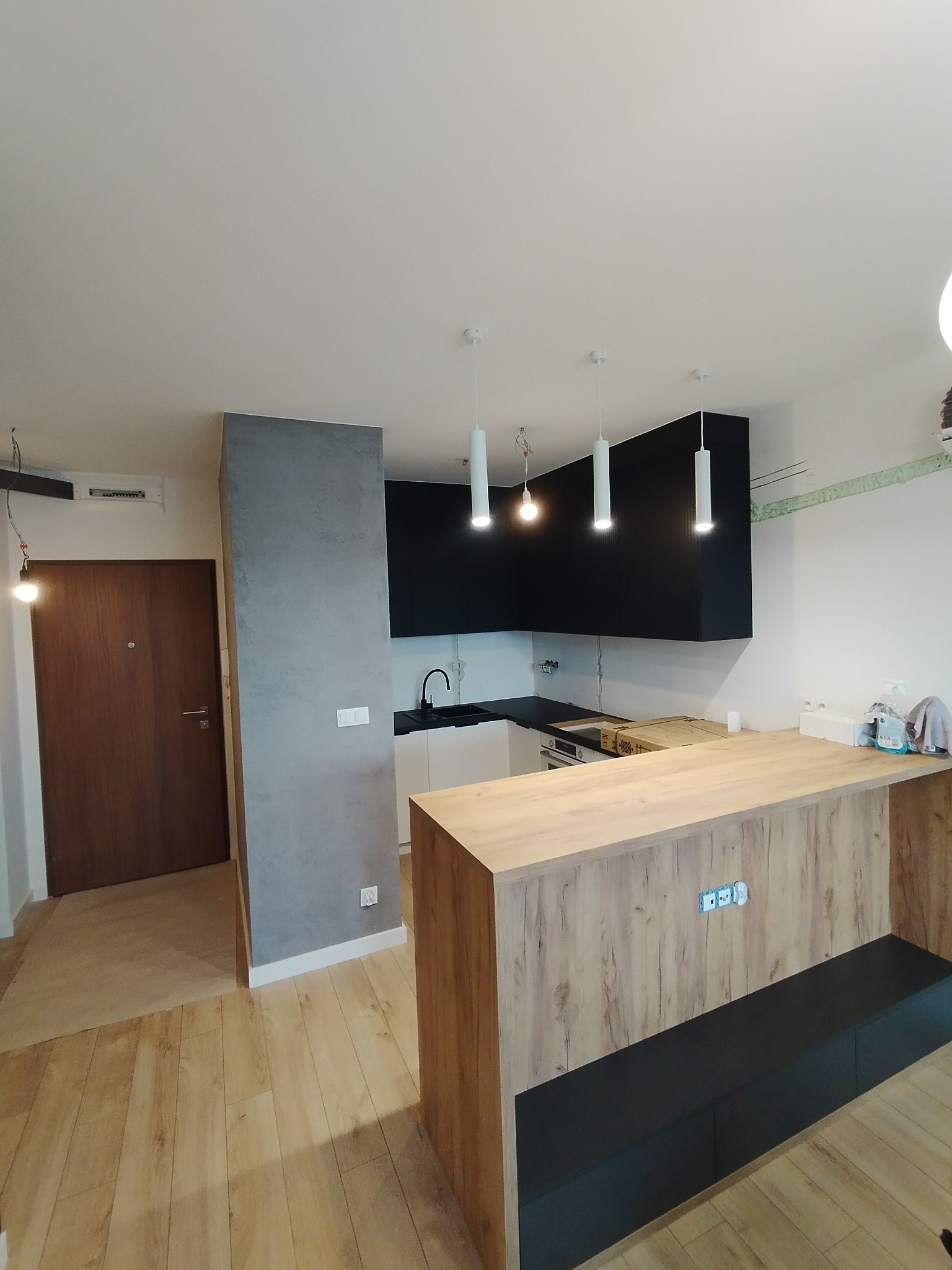 Nowoczesna kuchnia w Wasilkowie z drewnianą podłogą, szarą ścianą i czarnymi szafkami wiszącymi, oświetlona trzema cylindrycznymi lampami.