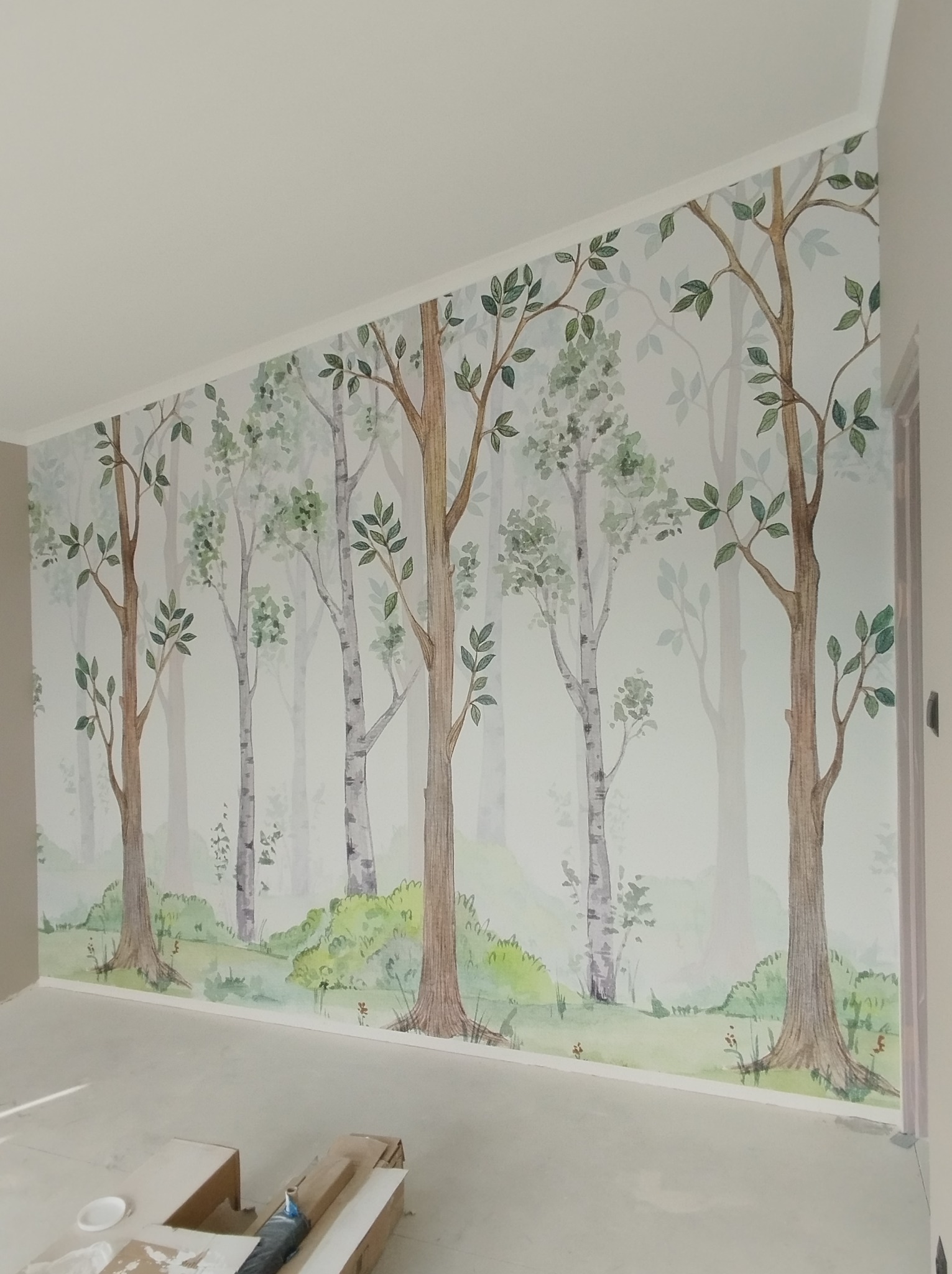 Ściana w pokoju dziecięcym z tapetą przedstawiającą akwarelowy las brzozowy z zielonym runem leśnym, widoczne narzędzia malarskie na podłodze.