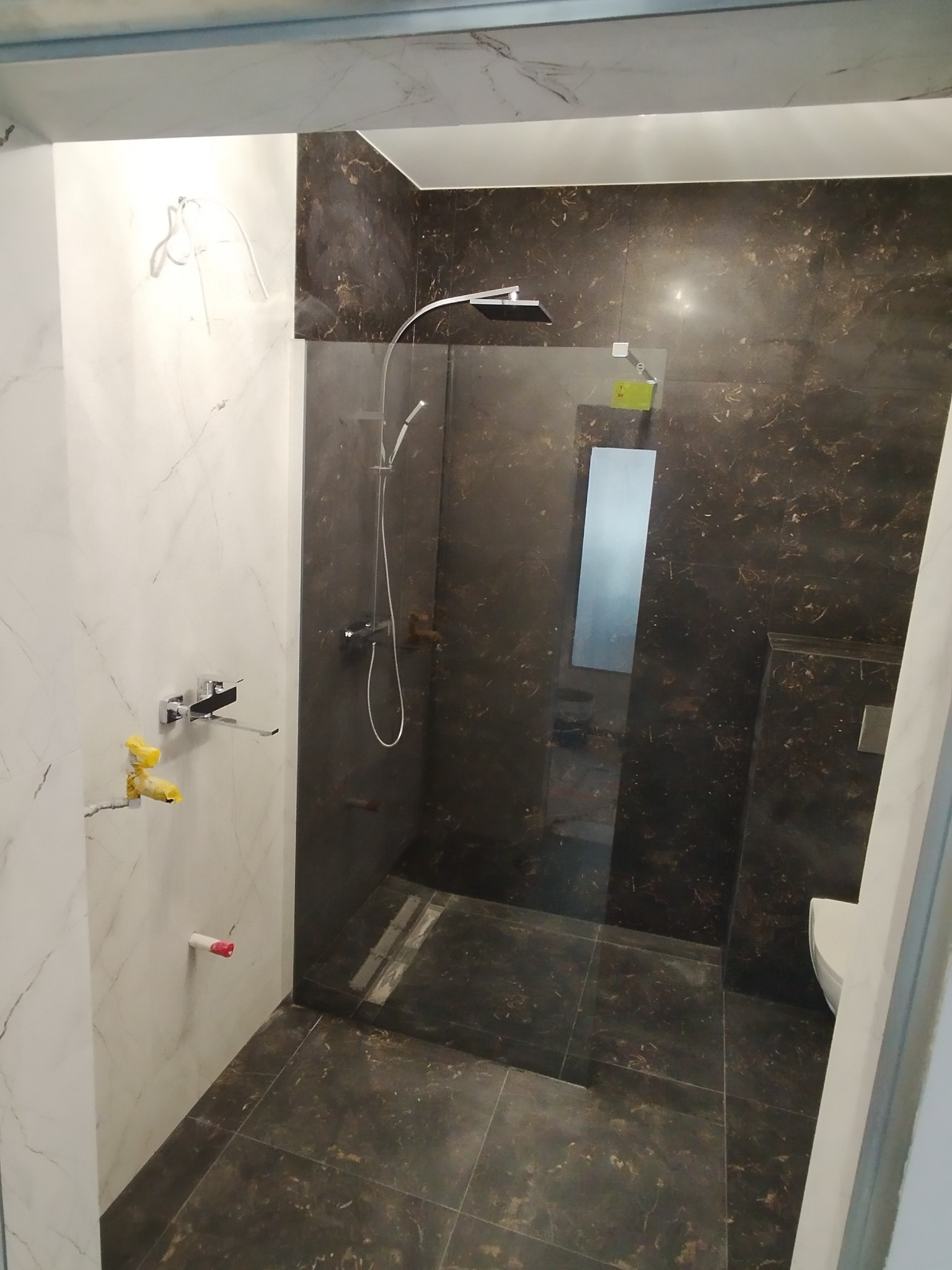 Nowoczesna kabina prysznicowa typu walk-in z panelami imitującymi marmur, widoczne przyłącza wodne zabezpieczone folią, biała ceramika sanitarna.