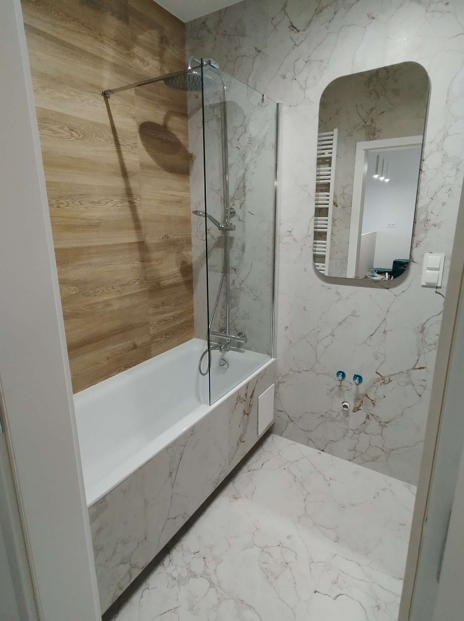 Nowoczesna łazienka z wanną obudowaną marmurem, przeszklonym parawanem nawannowym, lustrem o zaokrąglonych krawędziach i ścianami wykończonymi płytkami imitującymi marmur oraz drewno.