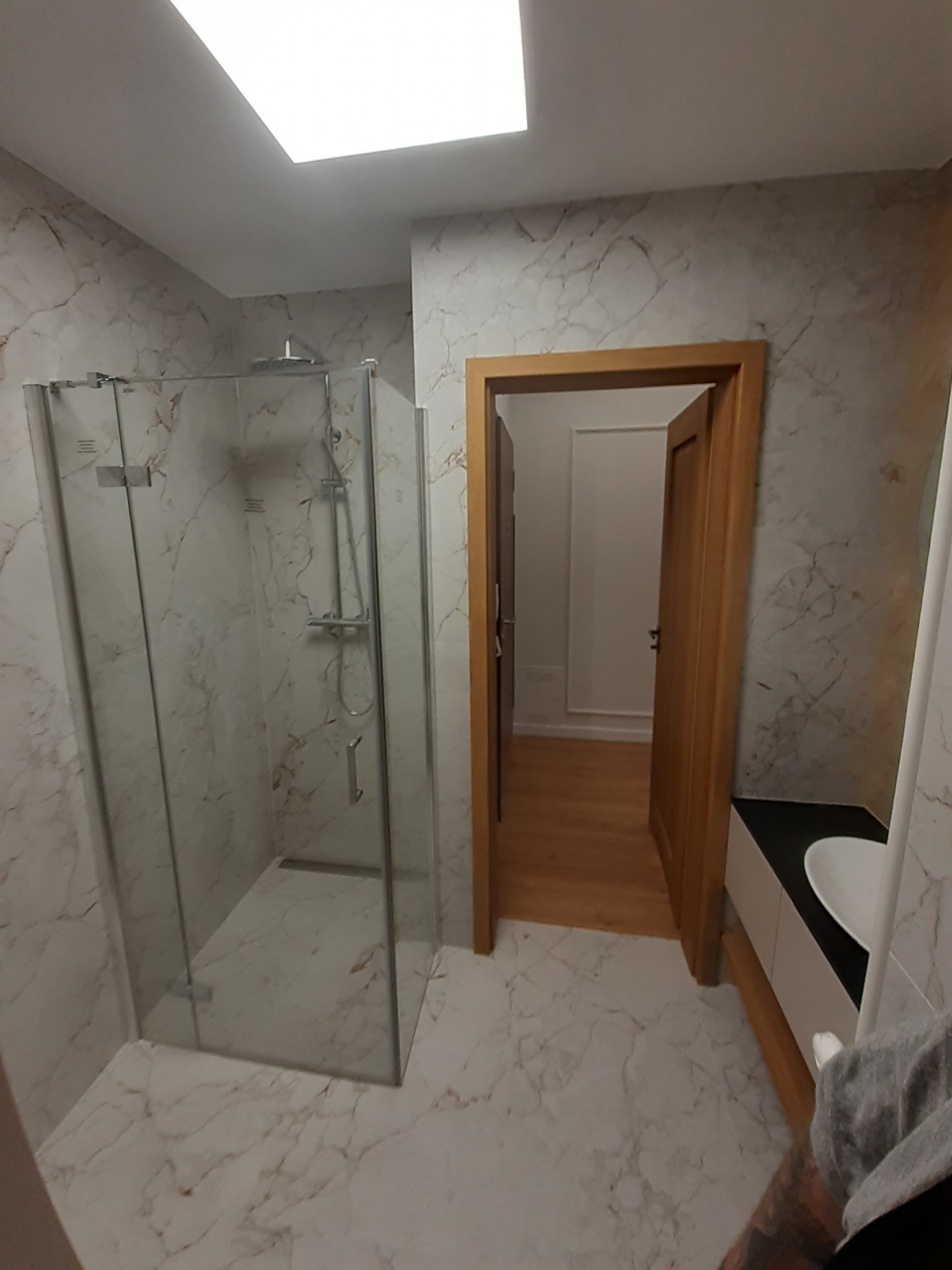 Nowoczesna łazienka z kabiną prysznicową typu walk-in, wykończona płytkami imitującymi marmur, z drewnianą ościeżnicą drzwiową i częściowo widoczną umywalką z szafką.
