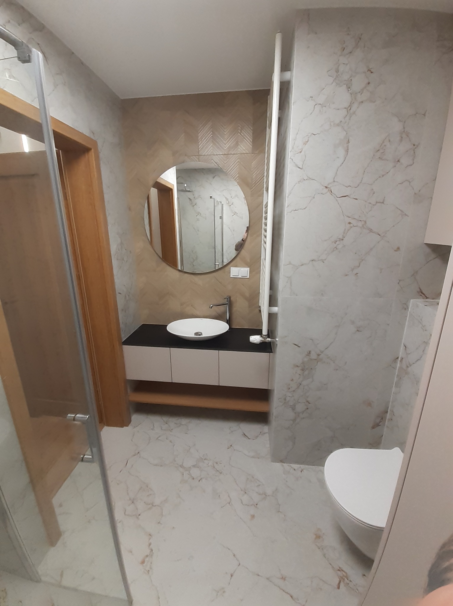 Nowoczesna łazienka z marmurowymi płytkami, okrągłym lustrem, białą umywalką nablatową na czarnym blacie i grzejnikiem drabinkowym.