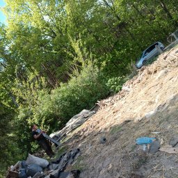 TRAW-KOP Tomasz Musiał - Nierówny teren ze stertą odpadów budowlanych i zielenią, mężczyzna trzymający narzędzie, w tle auto na pochyłości. Ujęcie z dołu, z widocznym datownikiem.