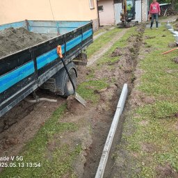 TRAW-KOP Tomasz Musiał - Wykop pod fundament z ułożonymi bloczkami betonowymi, na tle wywrotka z piaskiem i koparka. Widoczna osoba nadzorująca prace. Prace ziemne w toku.