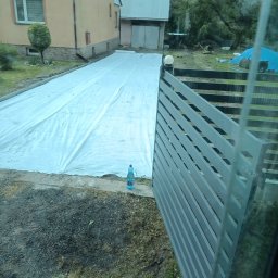 TRAW-KOP Tomasz Musiał - Przygotowanie terenu pod kostkę brukową, widoczne rozłożone płachty geowłókniny. W tle budynek mieszkalny i garaż. Nowoczesne ogrodzenie panelowe z metalu z boku.