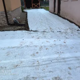 Rozłożona biała geowłóknina na ziemi wzdłuż budynku, widoczne ślady butów, obok budynku widać kostkę brukową i rynnę spustową. Przygotowanie terenu pod dalsze prace.