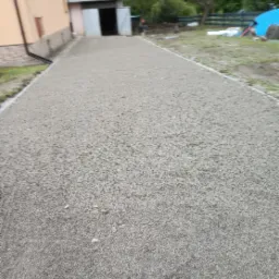 Ukończona podsypka z kruszywa pod nawierzchnię, z widocznym budynkiem gospodarczym w tle i fragmentem budynku mieszkalnego z rynną.