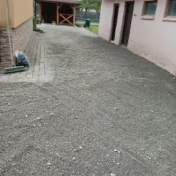 Ukończona ścieżka z szarego żwiru, prowadząca od kostki brukowej obok budynku do drewnianej altany w tle. Widoczny worek i miotła.