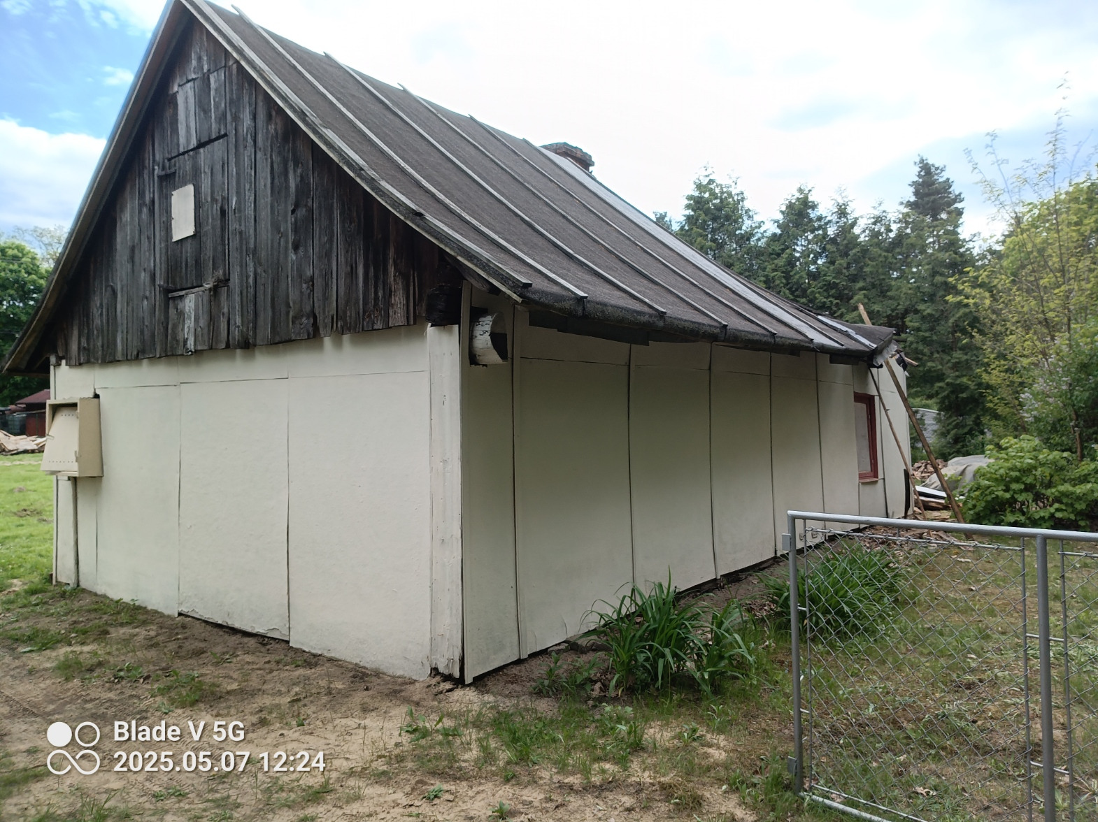 Stary, drewniany budynek z dobudowaną częścią, ogrodzony siatką. Widoczna zniszczona elewacja, drewniane elementy i fragment dachu. Otoczenie to trawa i drzewa.