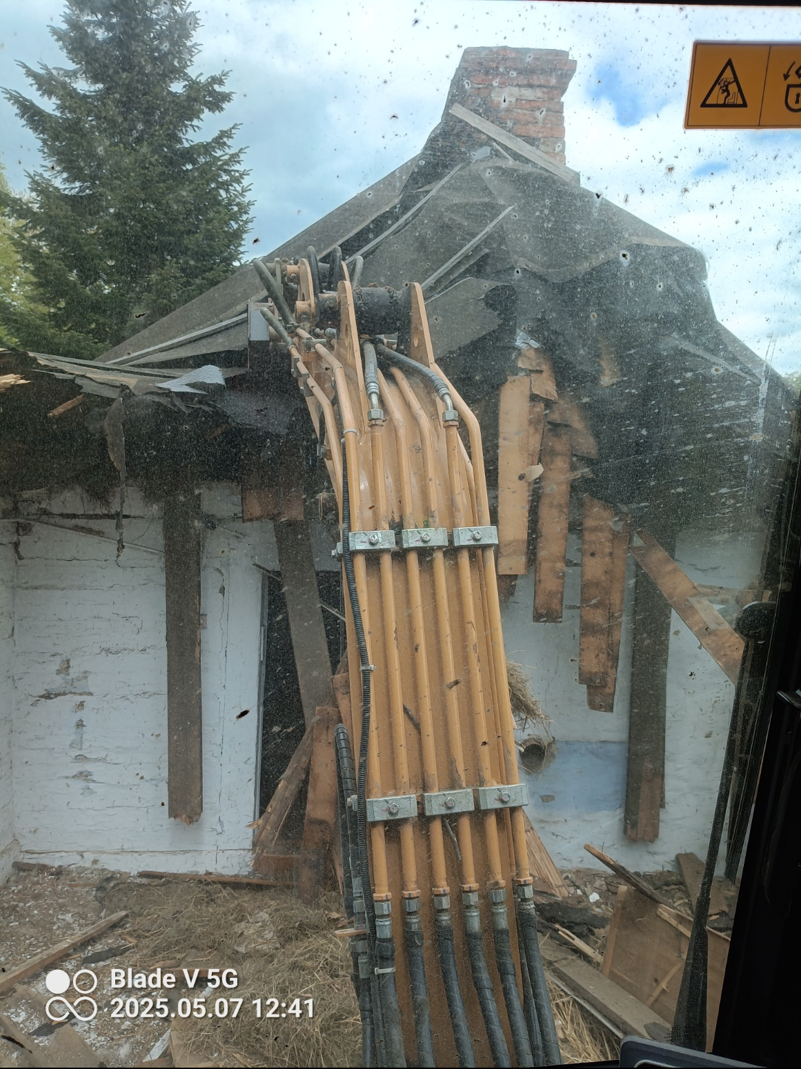 Ekskawator burzy budynek z ceglanym kominem. Widok z kabiny operatora, zarys drzewa w tle. Fragment dachu i ściany w trakcie rozbiórki.