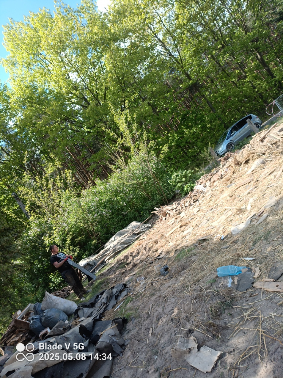 Nierówny teren ze stertą odpadów budowlanych i zielenią, mężczyzna trzymający narzędzie, w tle auto na pochyłości. Ujęcie z dołu, z widocznym datownikiem.