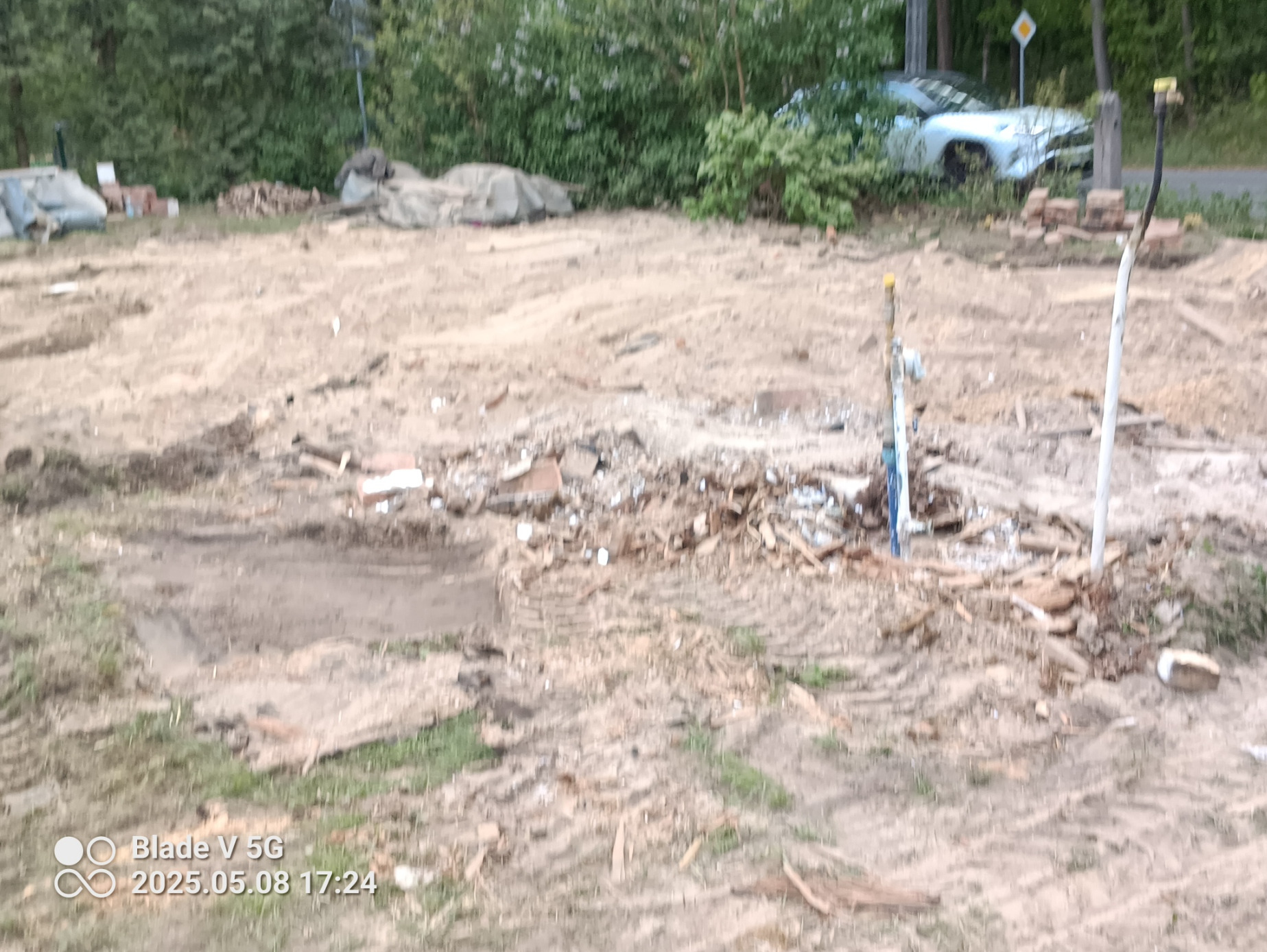 Teren po wyburzeniu z widocznymi śladami opon, gruzem i wystającymi rurami. W tle zieleń, samochód i znak drogowy. Fotografia z bliska.