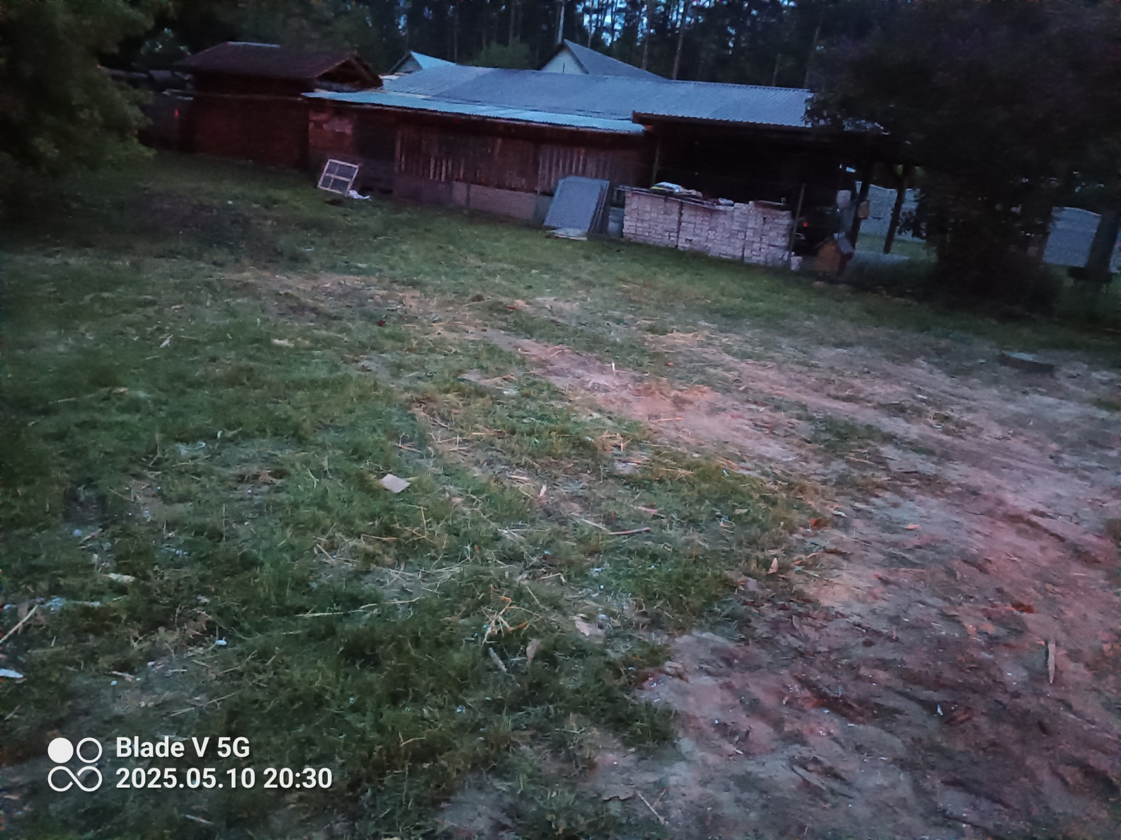 Teren z budynkami gospodarczymi w tle. Widoczne materiały budowlane, okno i trawa. Zdjęcie wykonane o zmierzchu, co wpływa na słabą widoczność detali.