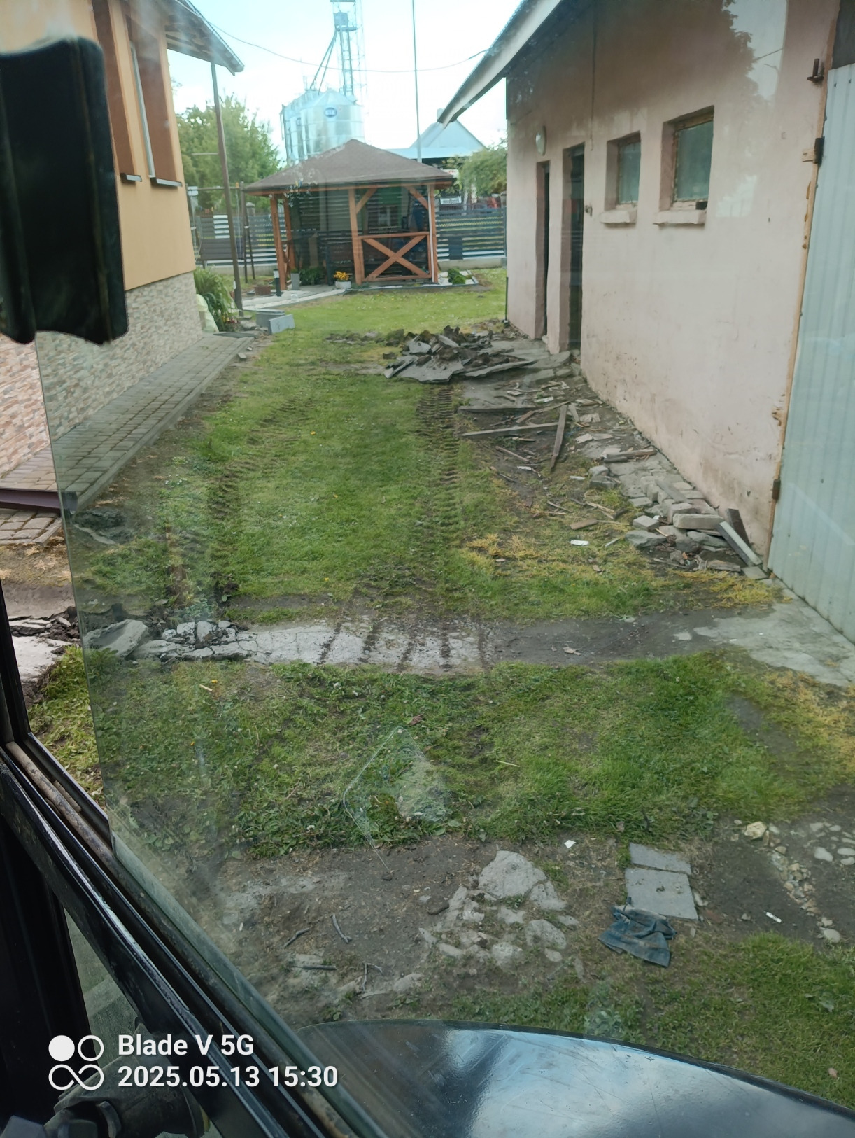 Teren po rozbiórce: ślady opon na trawie, gruz przy ścianie budynku, widok z kabiny maszyny. W tle altana i silos.