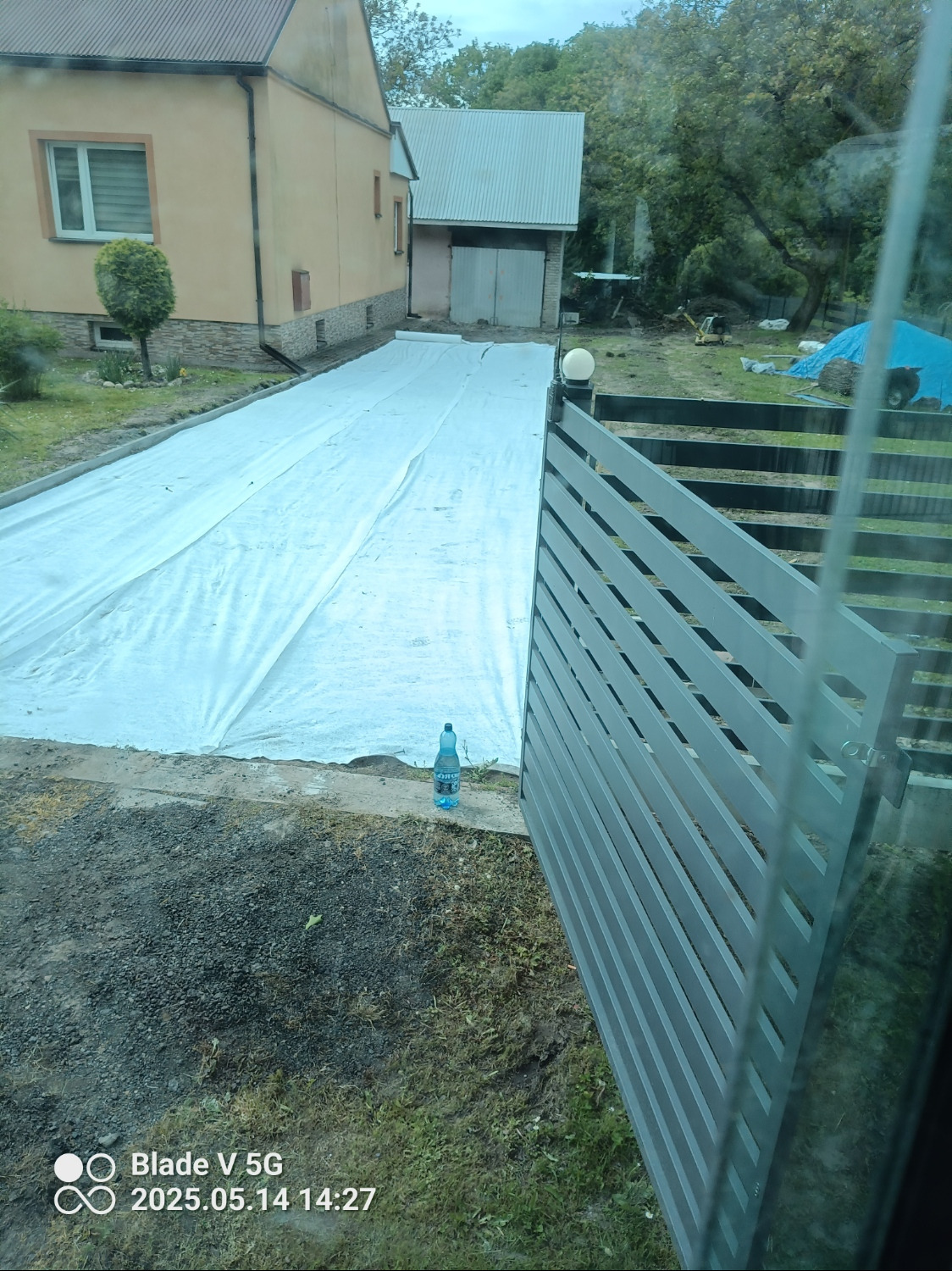 Przygotowanie terenu pod kostkę brukową, widoczne rozłożone płachty geowłókniny. W tle budynek mieszkalny i garaż. Nowoczesne ogrodzenie panelowe z metalu z boku.