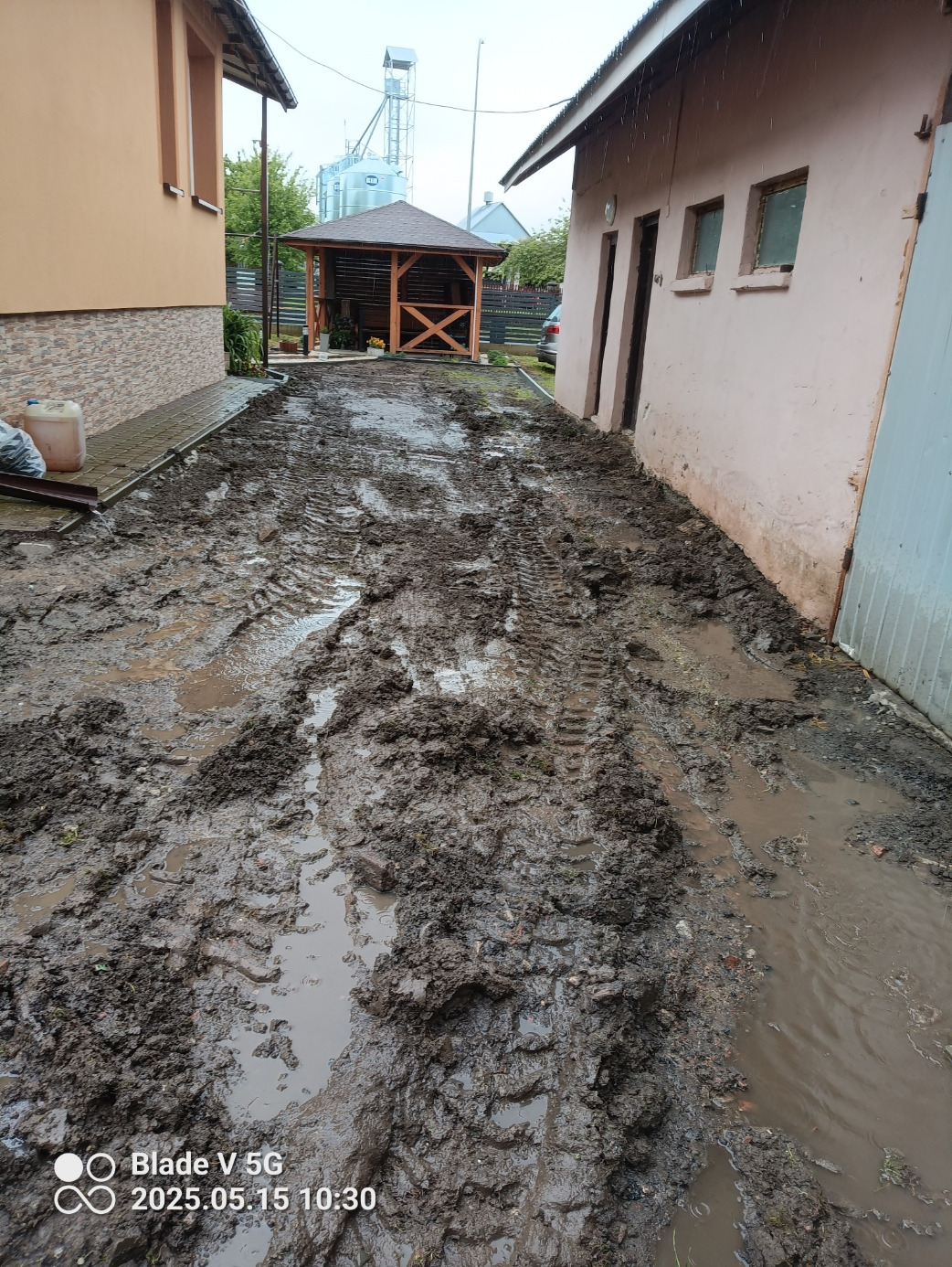 Błotnista droga z wyraźnymi śladami opon prowadząca do drewnianej altany, z budynkami po bokach. Ślady opon sugerują prace ziemne lub niwelację.