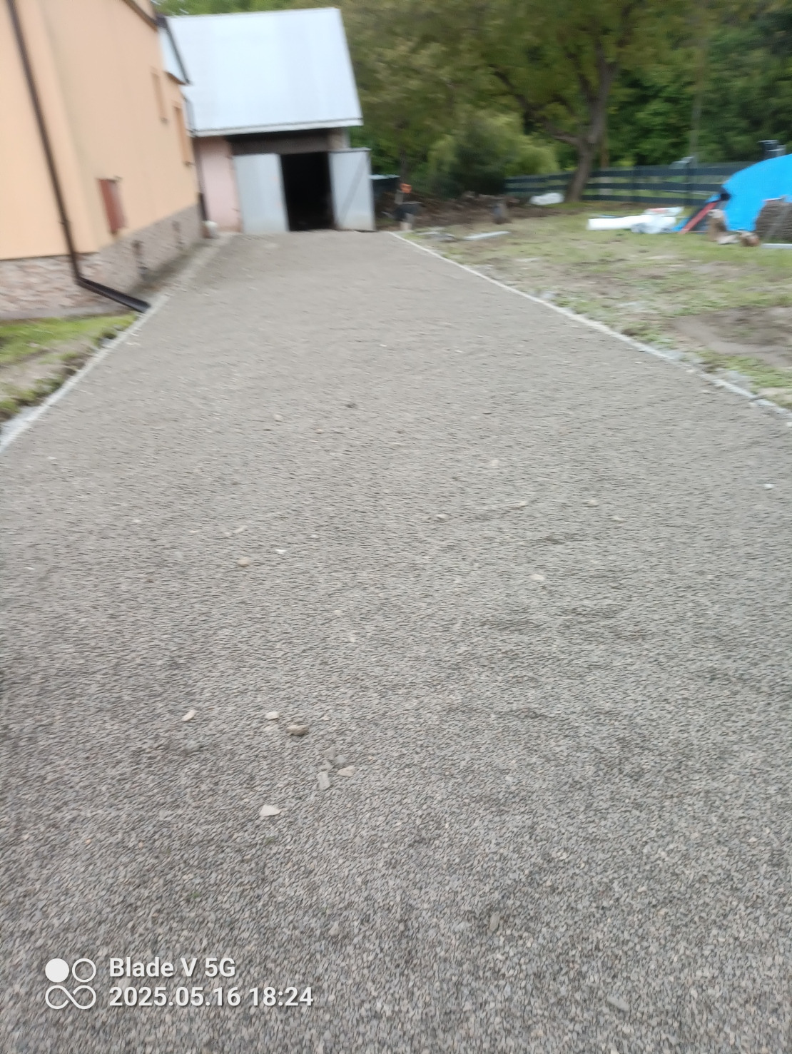 Ukończona podsypka z kruszywa pod nawierzchnię, z widocznym budynkiem gospodarczym w tle i fragmentem budynku mieszkalnego z rynną.