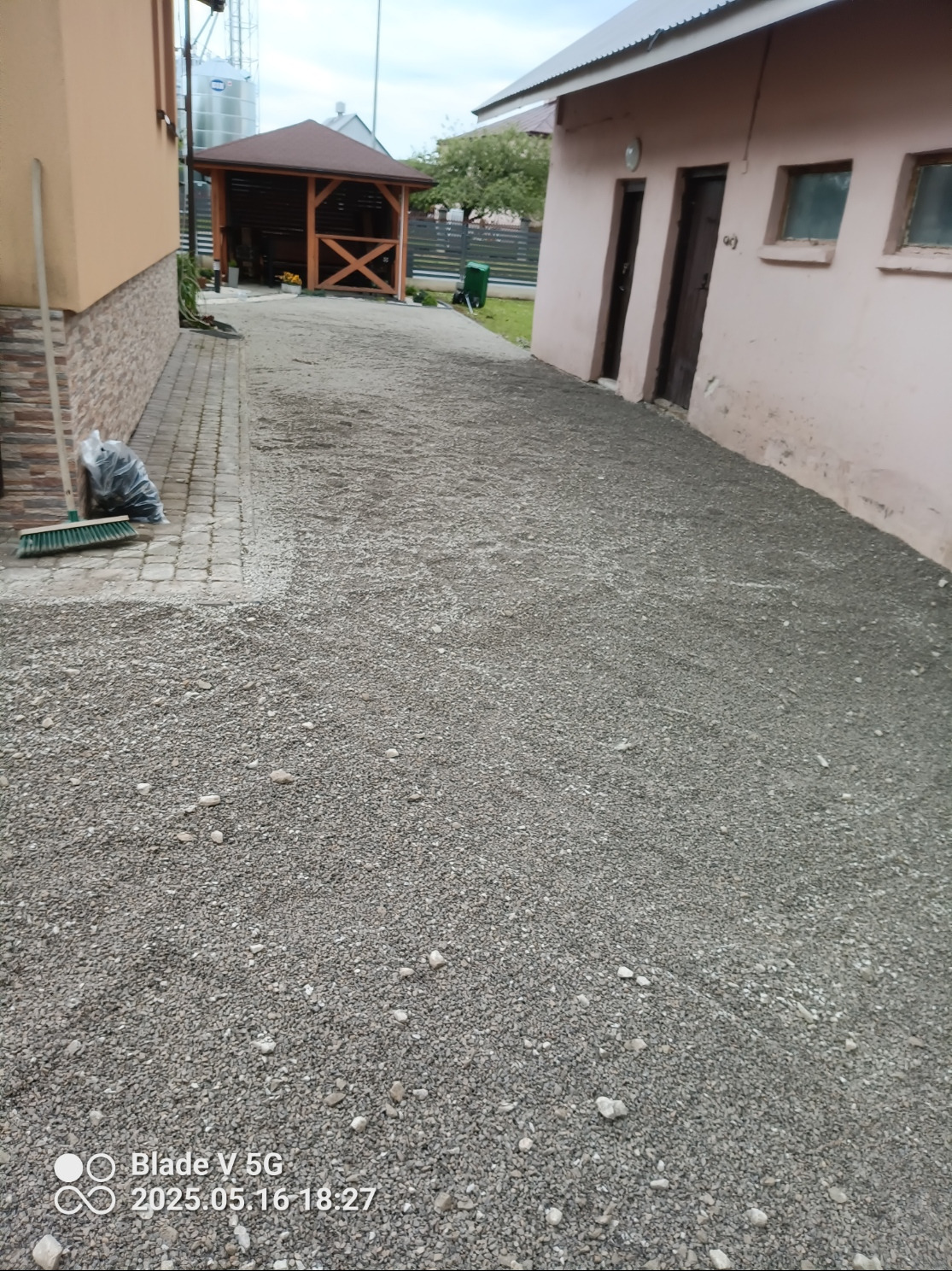 Ukończona ścieżka z szarego żwiru, prowadząca od kostki brukowej obok budynku do drewnianej altany w tle. Widoczny worek i miotła.