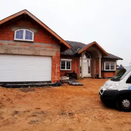 Nowo budowany dom z surowej cegły z zamontowaną białą bramą garażową segmentową i oknem łukowym, na posesji zaparkowany samochód dostawczy firmy budowlanej.