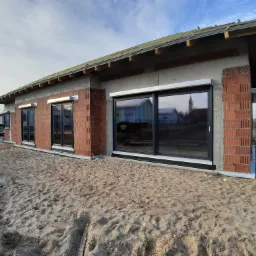 Surowy stan budowy domu jednorodzinnego z trzema dużymi oknami PCV z czarnymi ramami, widoczne cegły, beton, więźba dachowa i piasek na zewnątrz.