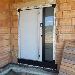 Montaż nowoczesnych drzwi wejściowych z długim, pionowym uchwytem, widoczne pianka montażowa i folia ochronna na ościeżnicy, surowy mur z cegły dziurawki w tle.