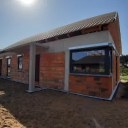 Budynek w stanie surowym z zamontowanymi oknami z czarną ramą i częściowo opuszczonymi roletami zewnętrznymi w kolorze srebrnym, widoczne cegły, betonowe elementy konstrukcyjne oraz piasek wokół...