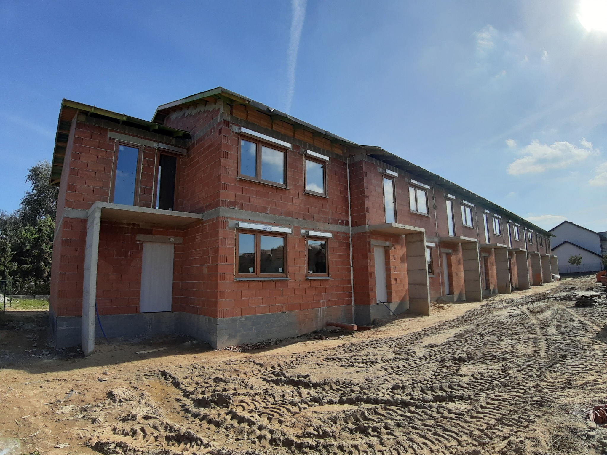 Surowy stan szeregowca z czerwonej cegły z zamontowanymi oknami z brązowymi ramami, widoczne ślady opon na placu budowy