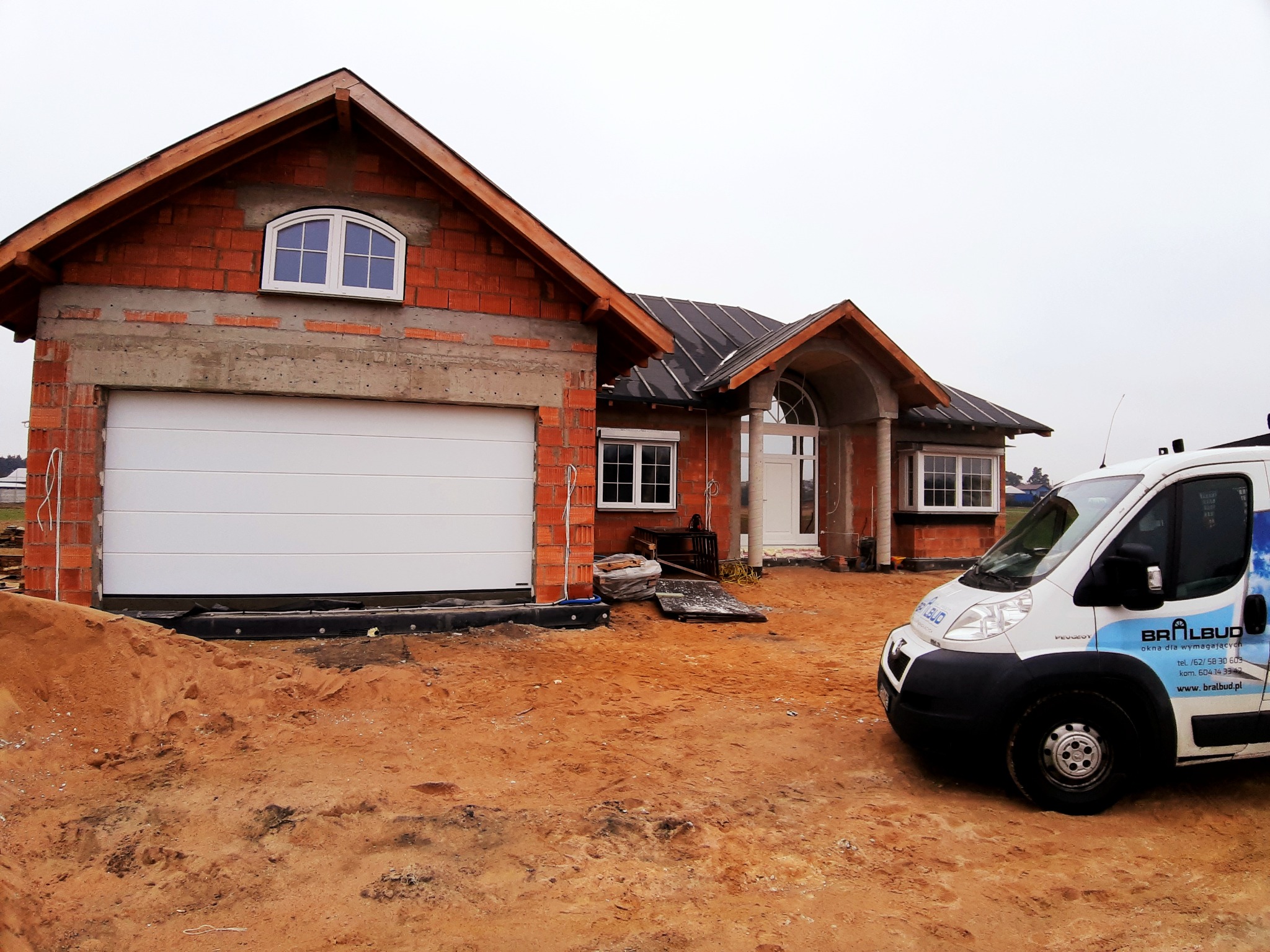 Nowo budowany dom z surowej cegły z zamontowaną białą bramą garażową segmentową i oknem łukowym, na posesji zaparkowany samochód dostawczy firmy budowlanej.