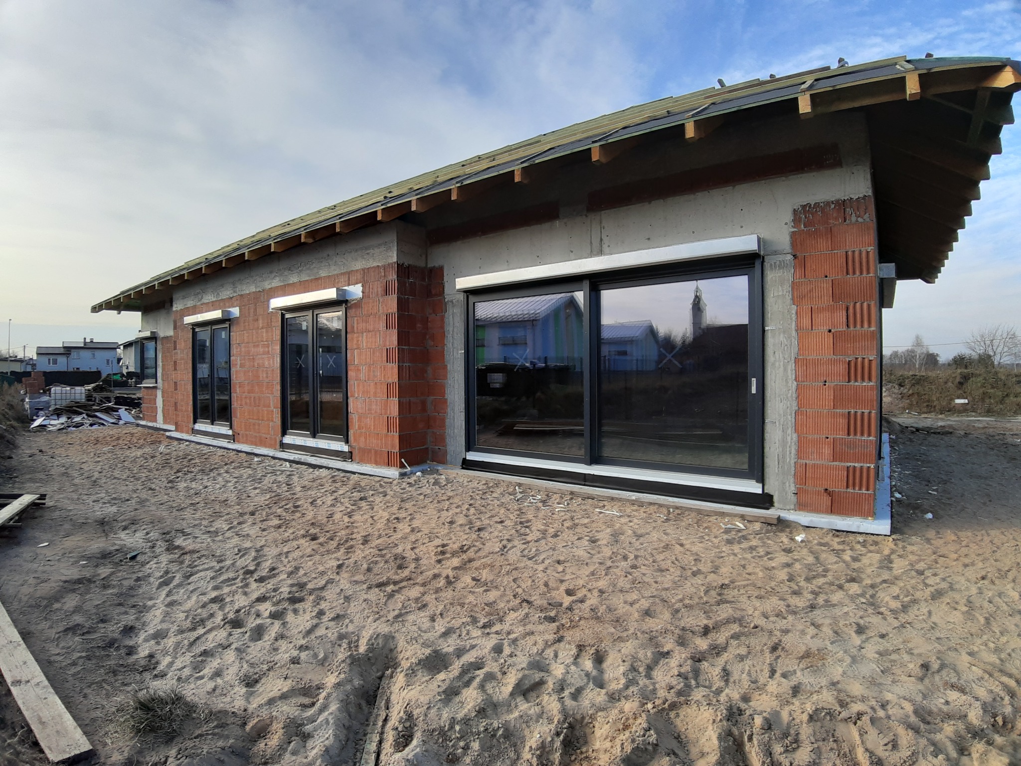 Surowy stan budowy domu jednorodzinnego z trzema dużymi oknami PCV z czarnymi ramami, widoczne cegły, beton, więźba dachowa i piasek na zewnątrz.