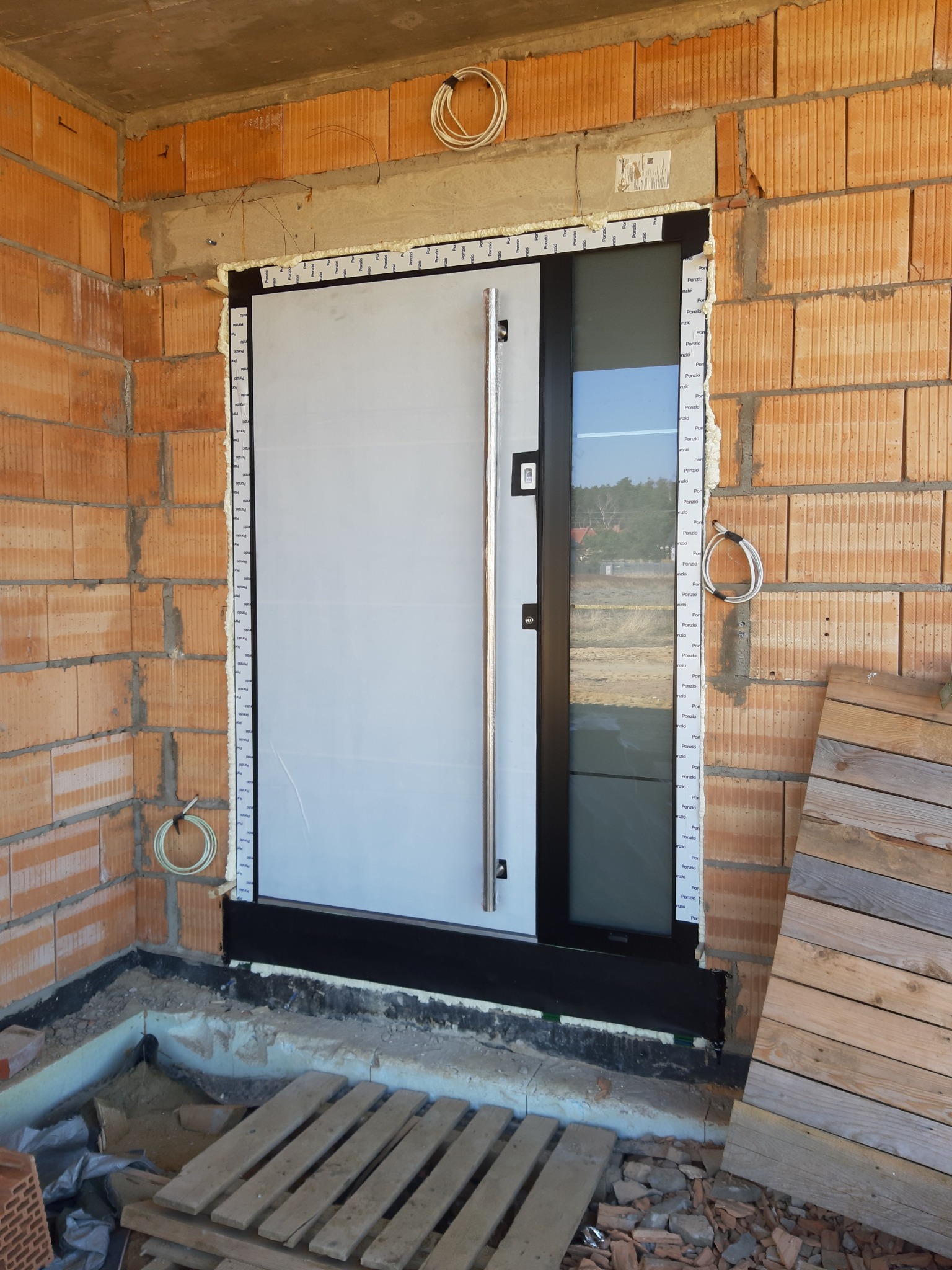 Montaż nowoczesnych drzwi wejściowych z długim, pionowym uchwytem, widoczne pianka montażowa i folia ochronna na ościeżnicy, surowy mur z cegły dziurawki w tle.