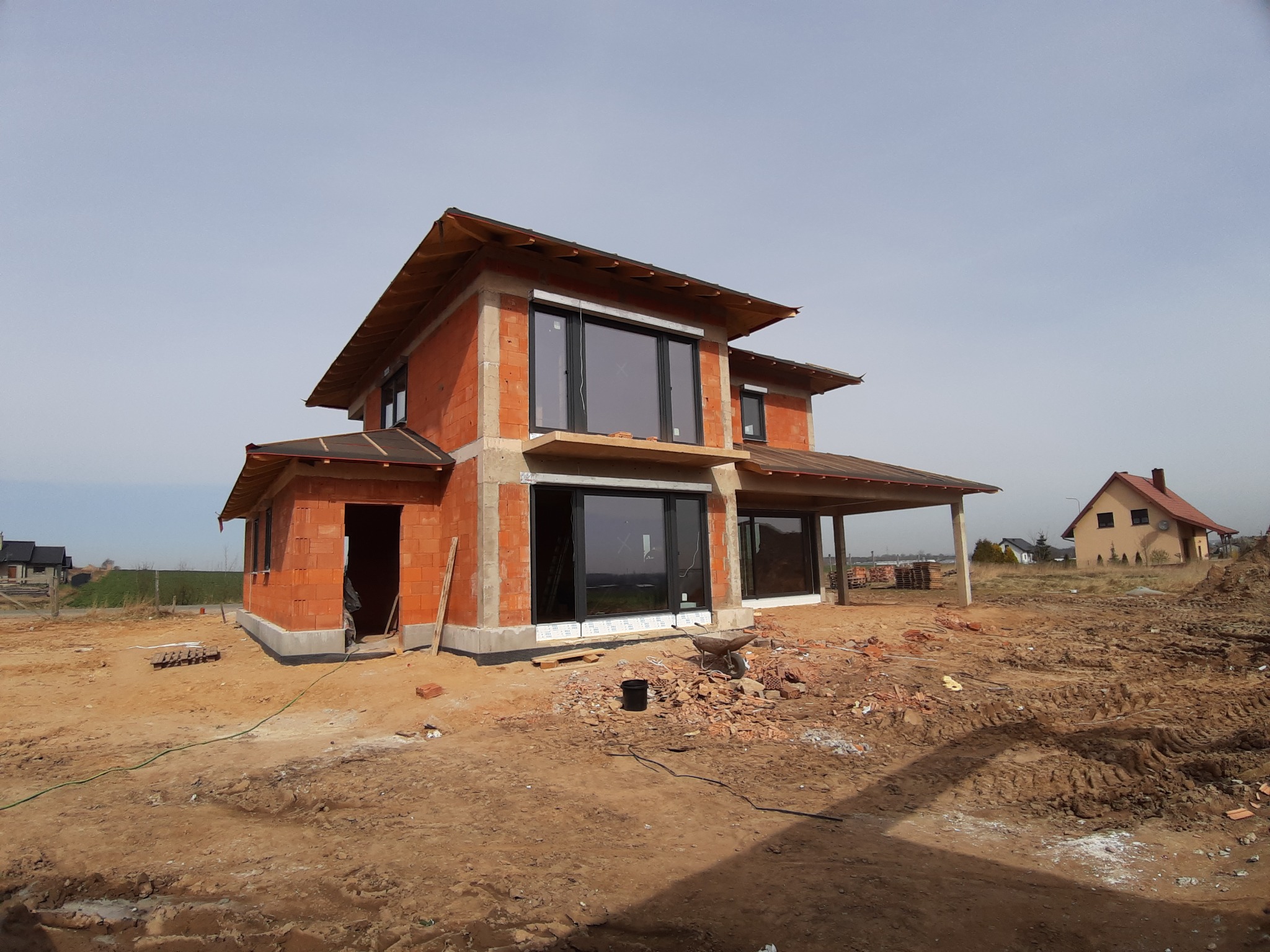 Dom w trakcie budowy z widocznymi oknami z czarnymi ramami, surowy stan, cegła, przygotowany teren.