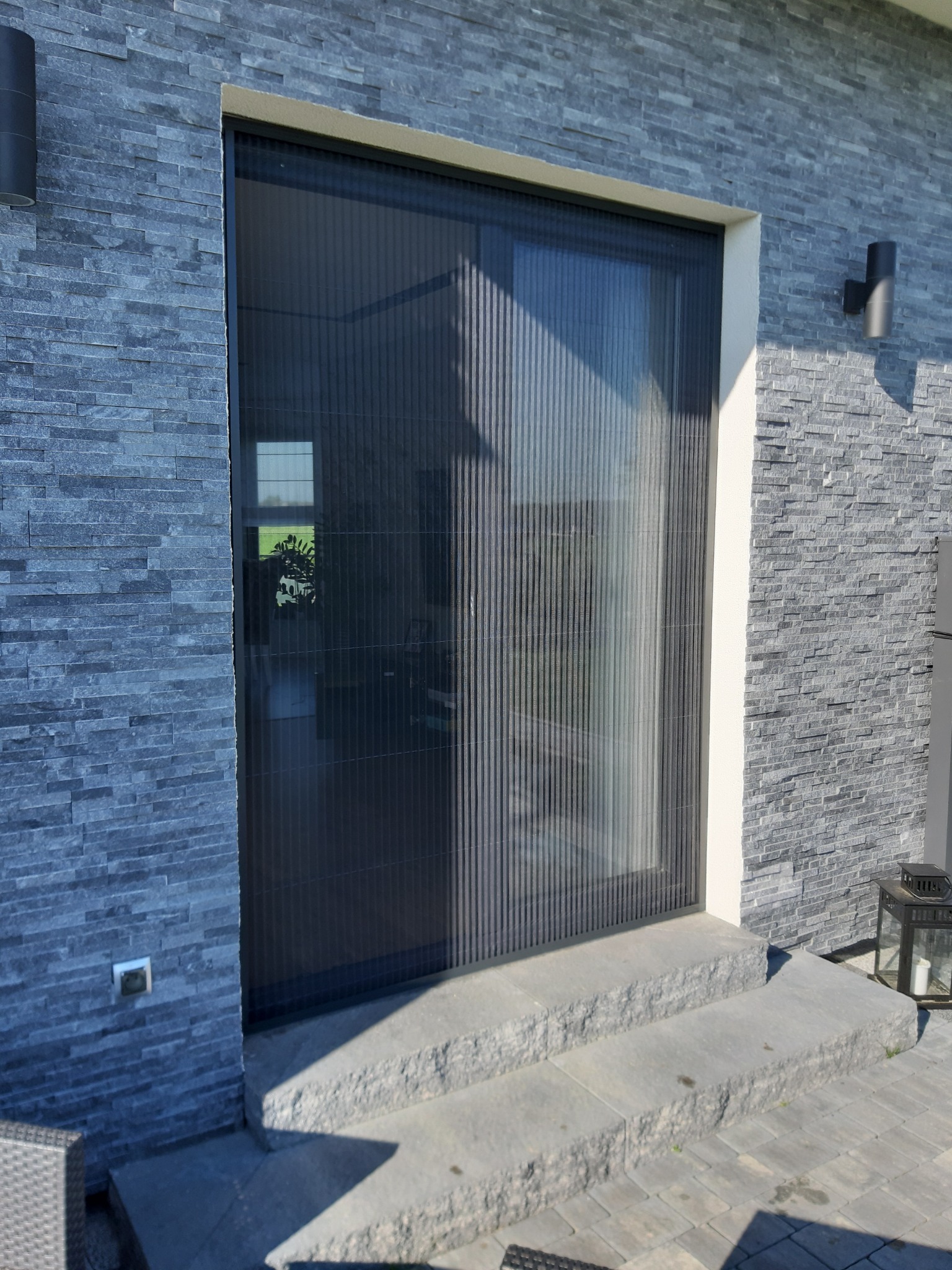 Zewnętrzne przesuwne drzwi z moskitierą plisowaną, zamontowane w elewacji z szarej, łupanej płytki kamiennej, widoczne trzy kamienne stopnie wejściowe i lampa elewacyjna.