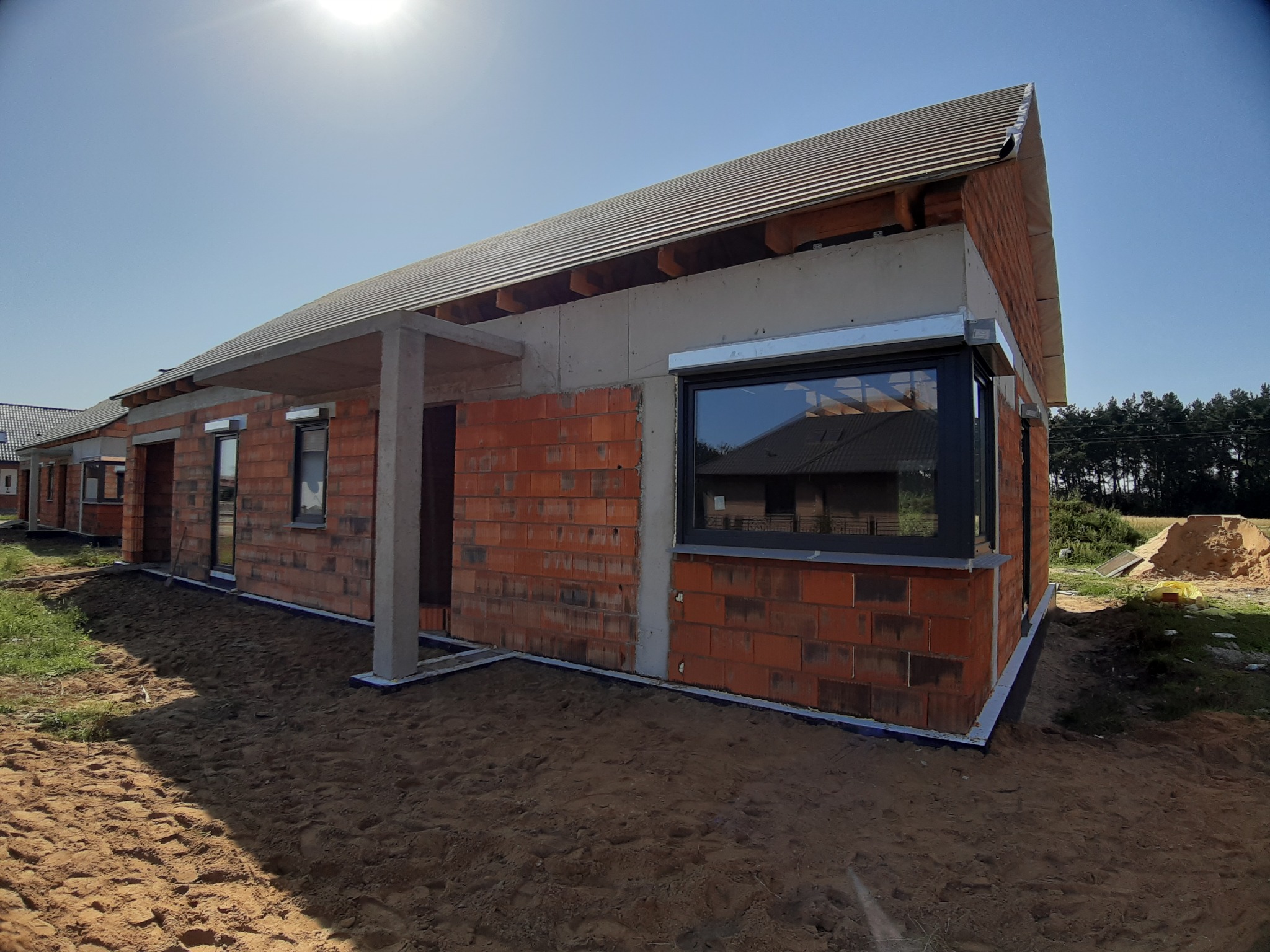 Budynek w stanie surowym z zamontowanymi oknami z czarną ramą i częściowo opuszczonymi roletami zewnętrznymi w kolorze srebrnym, widoczne cegły, betonowe elementy konstrukcyjne oraz piasek wokół...